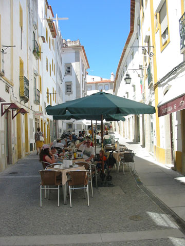 Patrons sitting at shaded tables outside a street café along a historic street in Évora, Alentejo